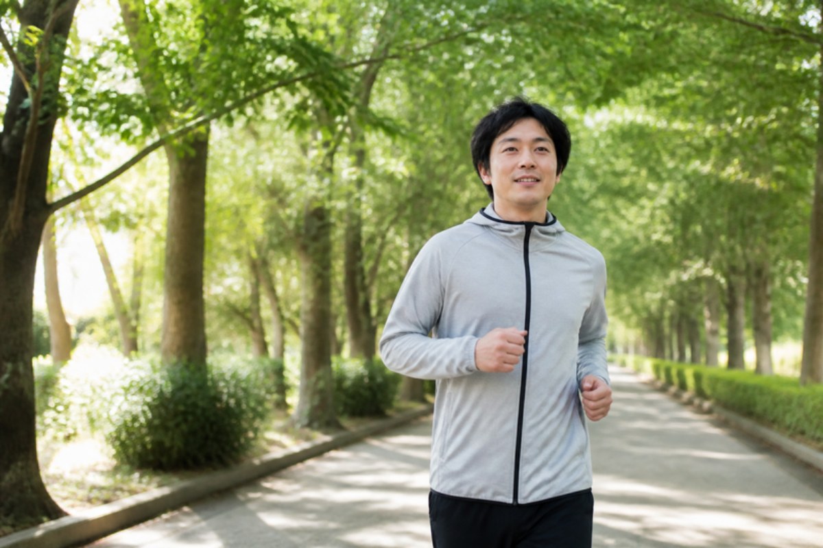 朝の光が差す並木道の公園を、軽い運動着で、ほほえみながら歩く男性の写真