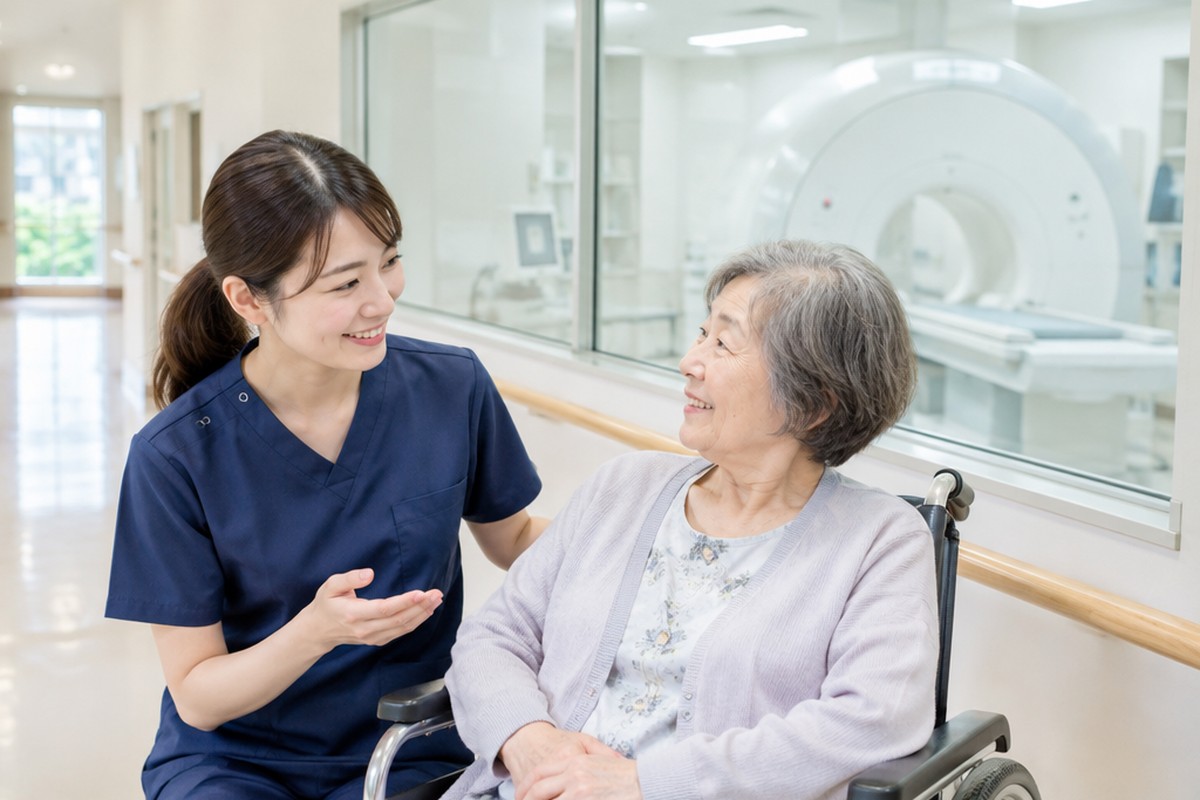 病院のMRI室の前で、看護師が車いすの高齢の女性に笑顔で説明している